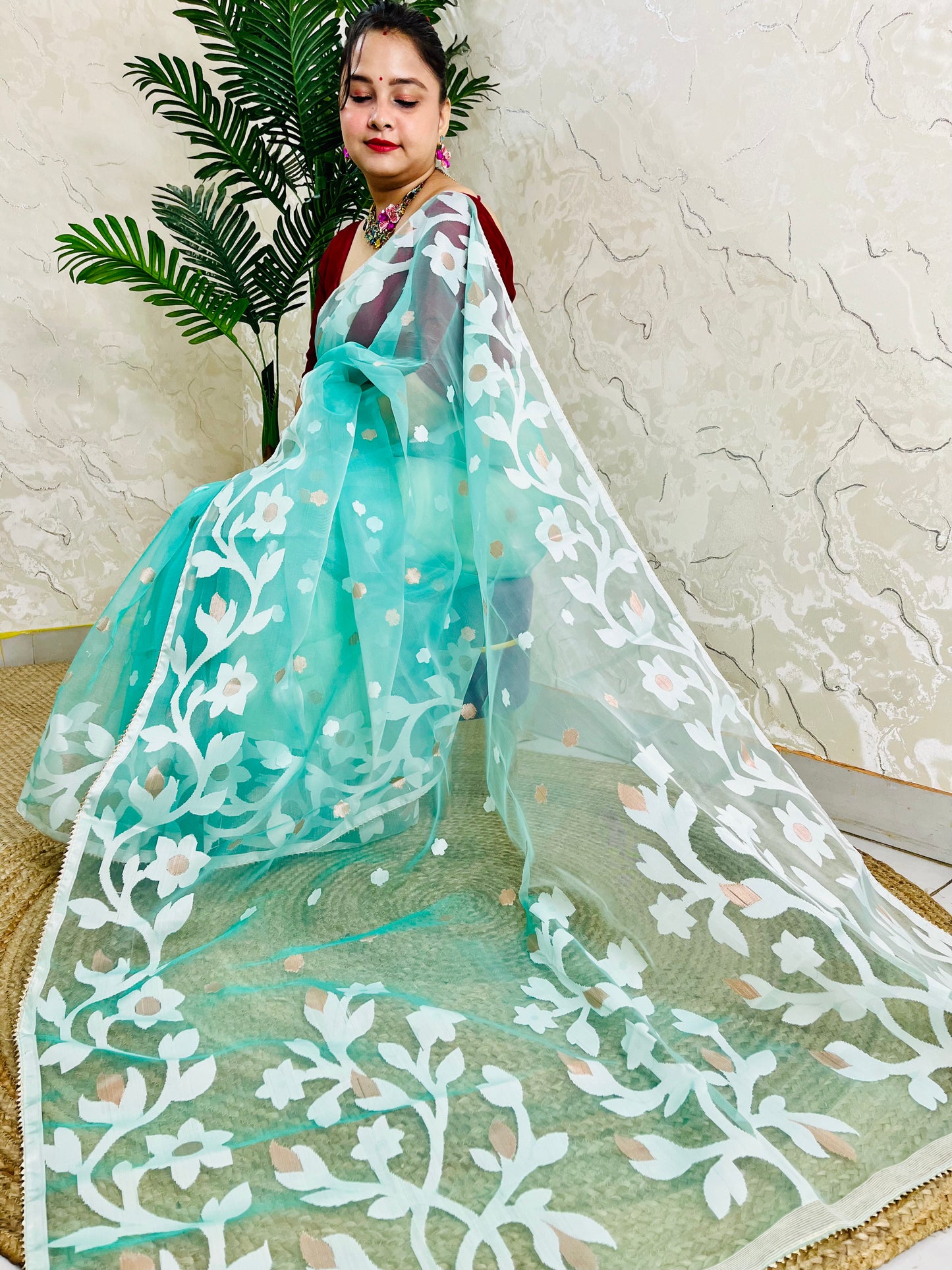 A woman wearing a light teal-green saree with white floral embroidery and a matching dupatta, posed in front of a textured beige wall with a potted plant.