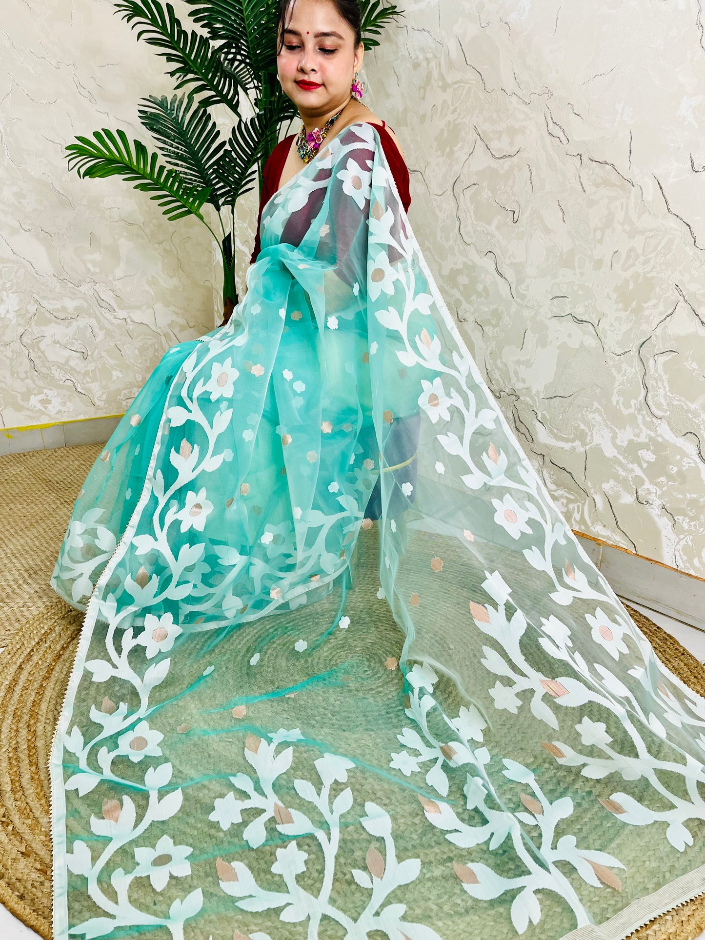 A woman wearing a light turquoise blue saree with white floral embroidery and a matching dupatta, posed in front of a textured beige wall with a green plant in the background.