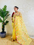 A woman wearing a yellow saree with colorful floral motifs and a red blouse, standing indoors next to a potted palm tree against a textured beige wall.
