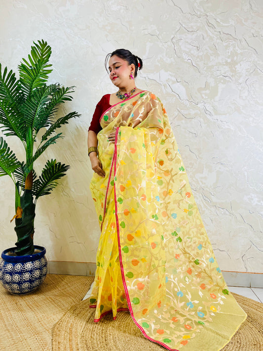 A woman wearing a yellow saree with colorful floral motifs and a red blouse, standing indoors next to a potted palm tree against a textured beige wall.