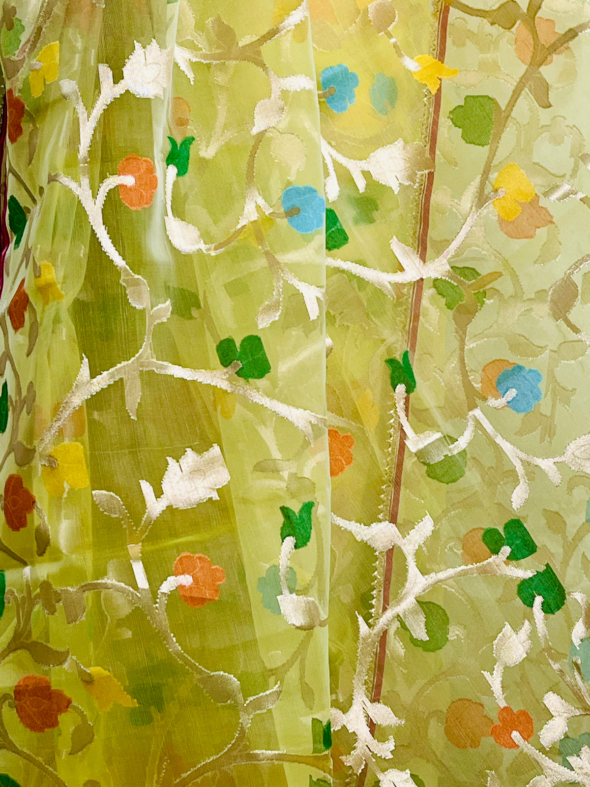 A close-up of a sheer light green fabric with embroidered white floral and vine patterns, accented by small painted flowers in blue, orange, and yellow.