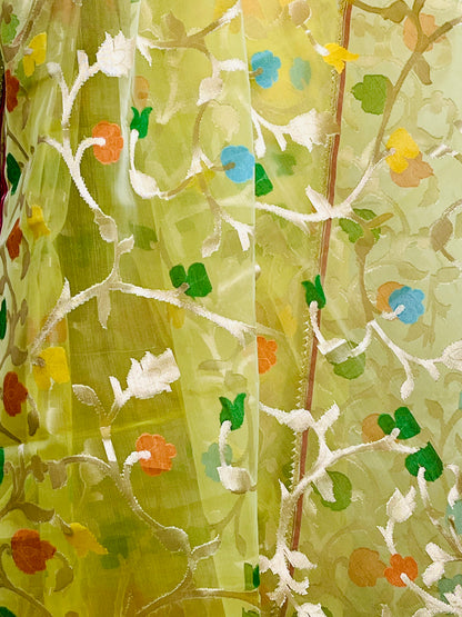 A close-up of a sheer light green fabric with embroidered white floral and vine patterns, accented by small painted flowers in blue, orange, and yellow.