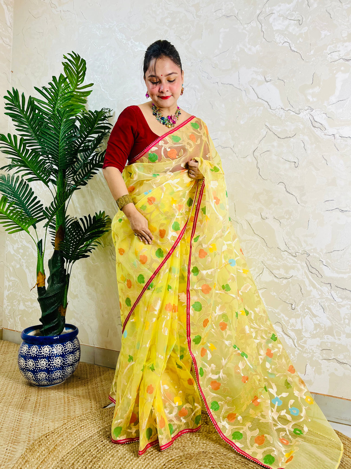 A woman wearing a yellow saree with colorful floral prints and a red blouse, standing indoors next to a potted palm tree against a textured beige wall.