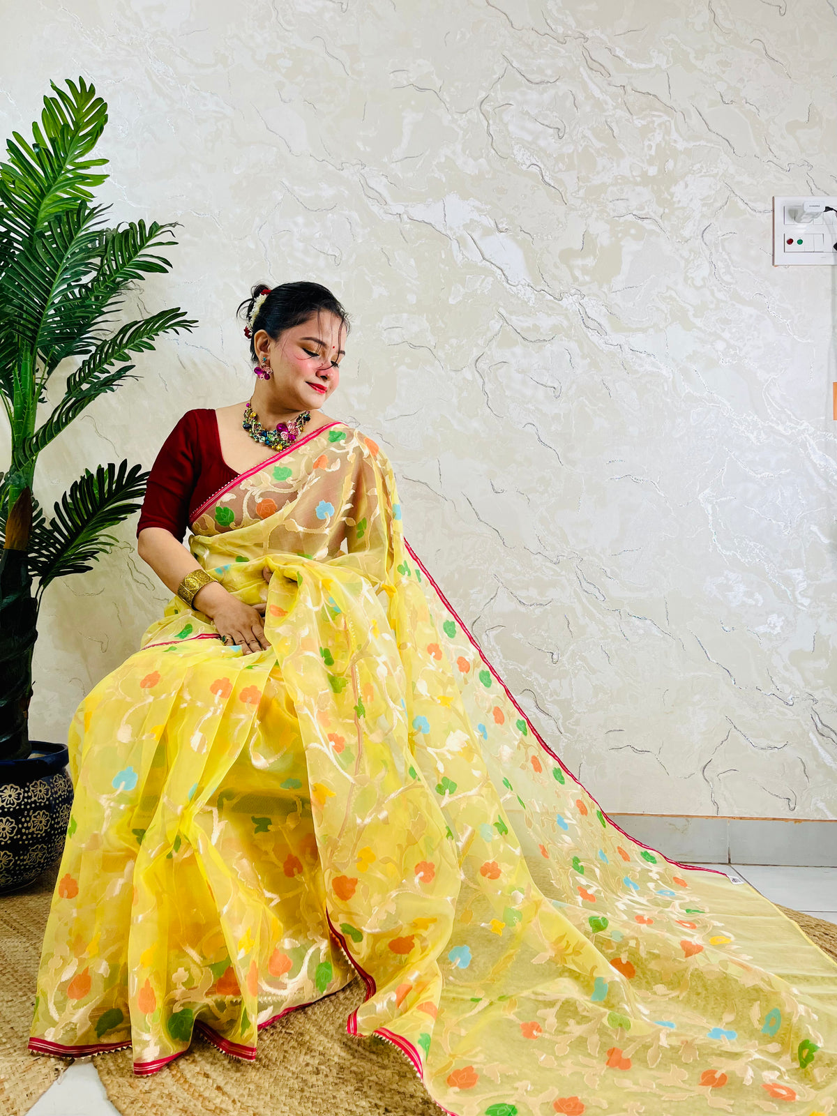 A woman wearing a yellow saree with colorful floral patterns and a deep red blouse, seated indoors against a textured cream wall next to a potted plant.