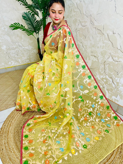 A woman wearing a yellow embroidered saree with multicolored floral motifs and a red border, seated on a woven mat against a textured beige wall with a green plant in the background.