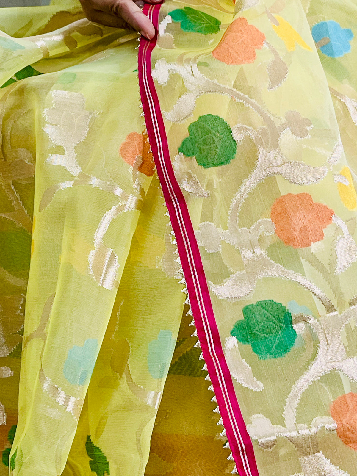 A close-up of a light yellow saree with white floral motifs and colorful embroidered floral accents in green, orange, and blue. A hand is holding the edge of the saree, which features a bright pink border with white stripes and a serrated edge.