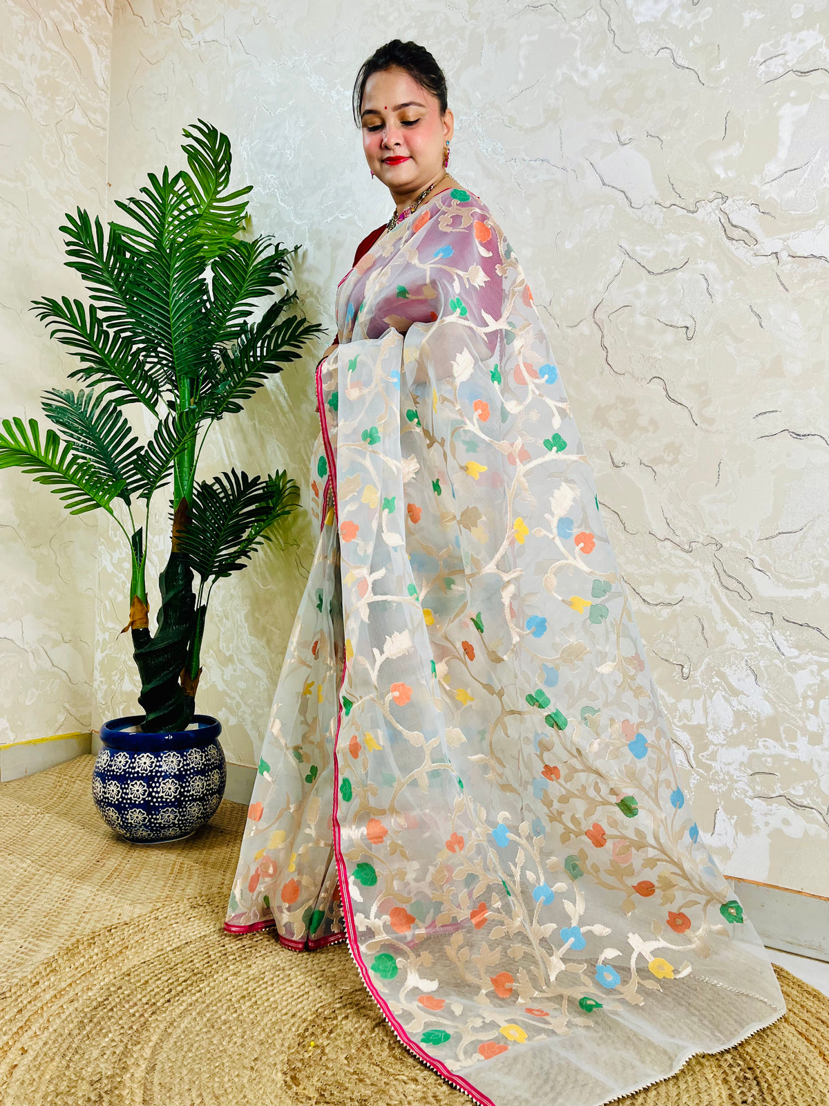 A woman wearing a white organza saree with colorful floral embroidery and a pink border, standing next to a potted palm tree in front of a textured beige wall.