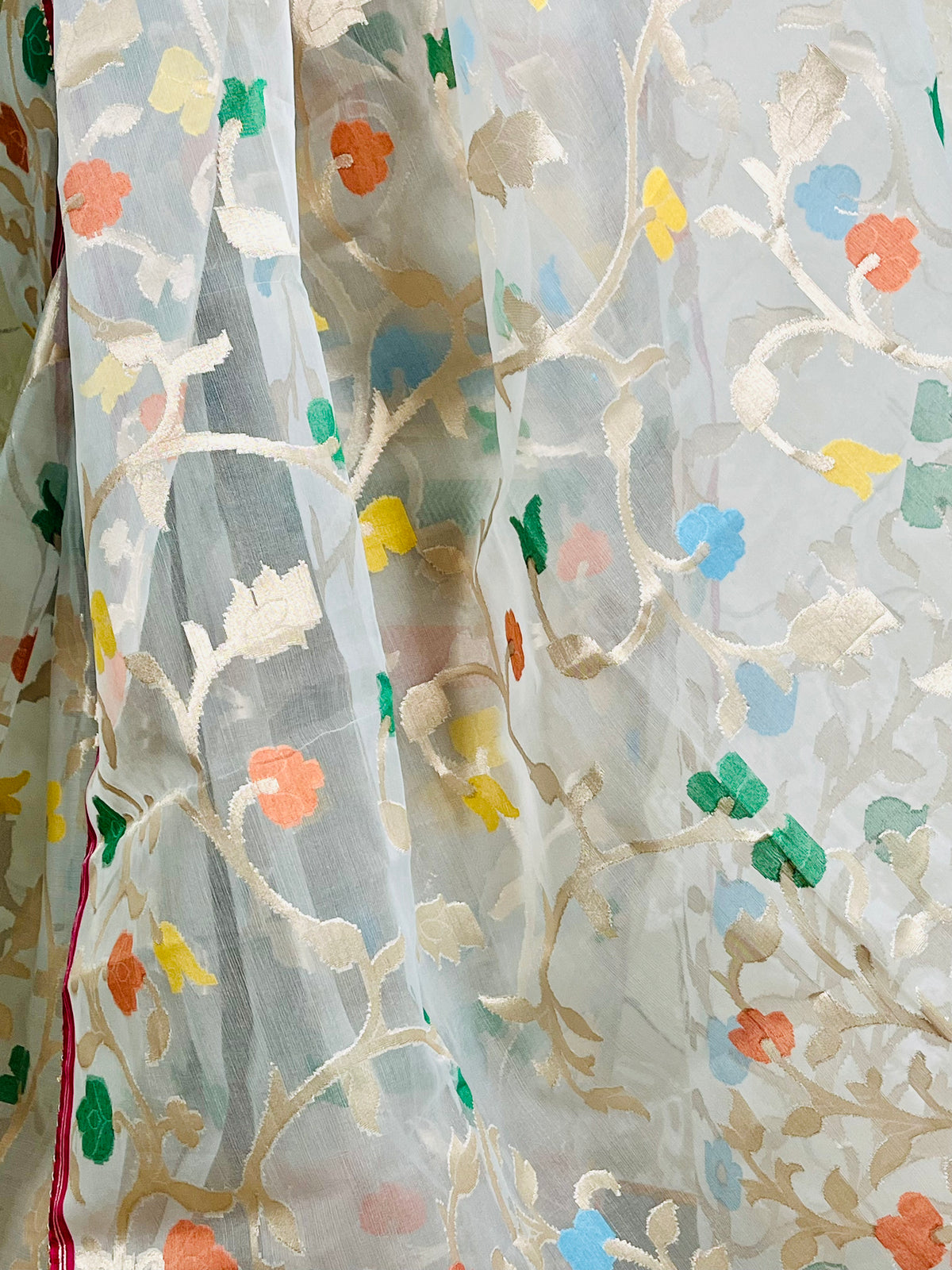 A close-up of a sheer white organza fabric with intricate gold floral embroidery and scattered colorful floral accents in orange, blue, green, and yellow.
