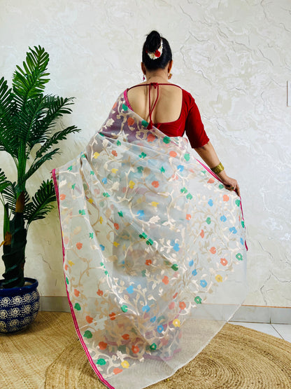 A woman viewed from behind wearing a white saree with colorful floral embroidery and a red blouse, standing indoors next to a potted plant against a textured wall.