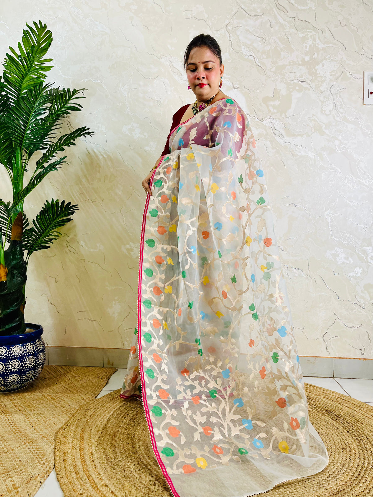 A woman models a white organza saree with colorful floral embroidery and a pink border, standing in a room with a textured beige wall, a potted plant, and a woven mat.