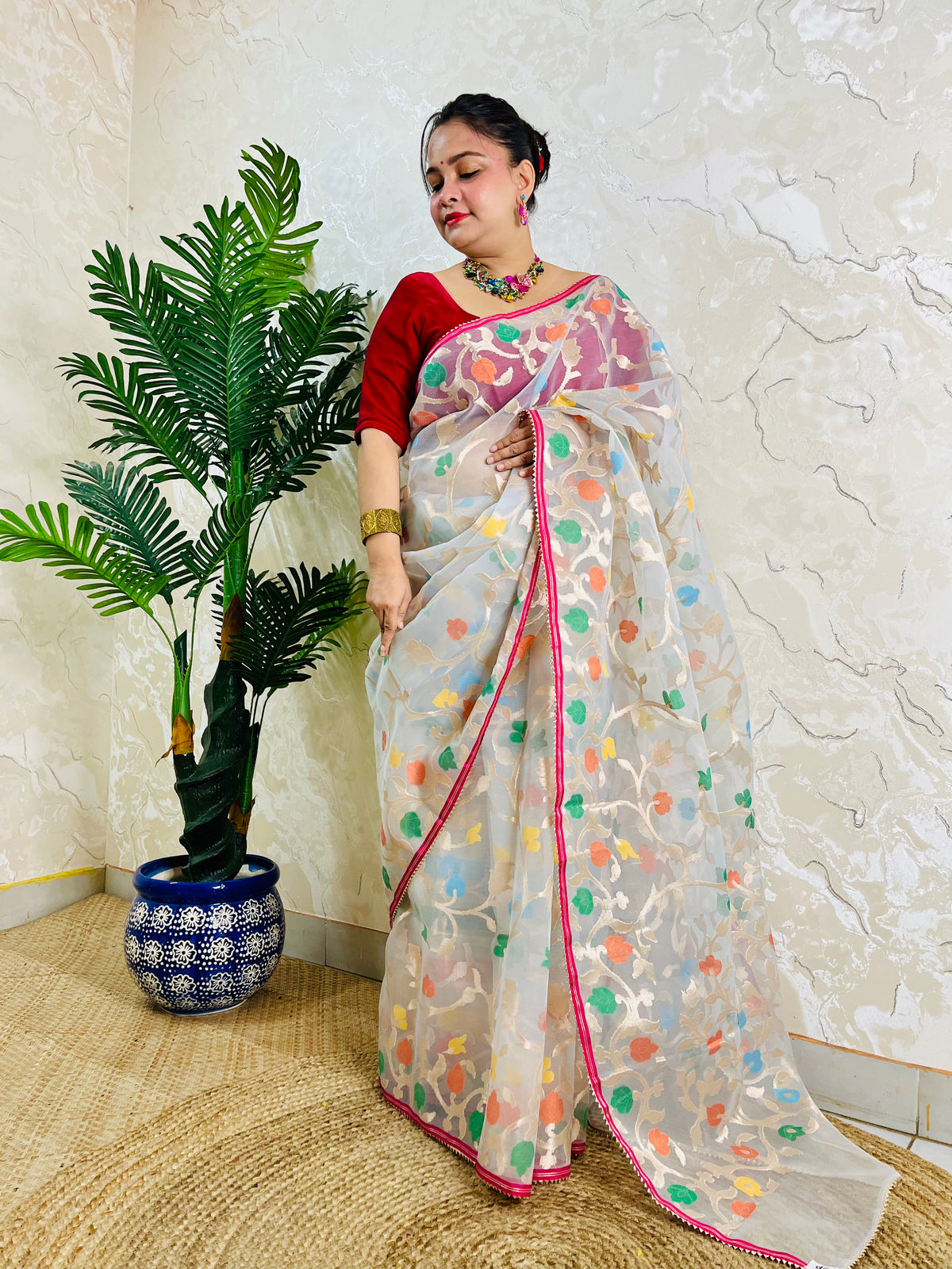 A woman wearing a white saree with colorful floral prints and a red blouse, standing indoors next to a potted plant against a textured wall.