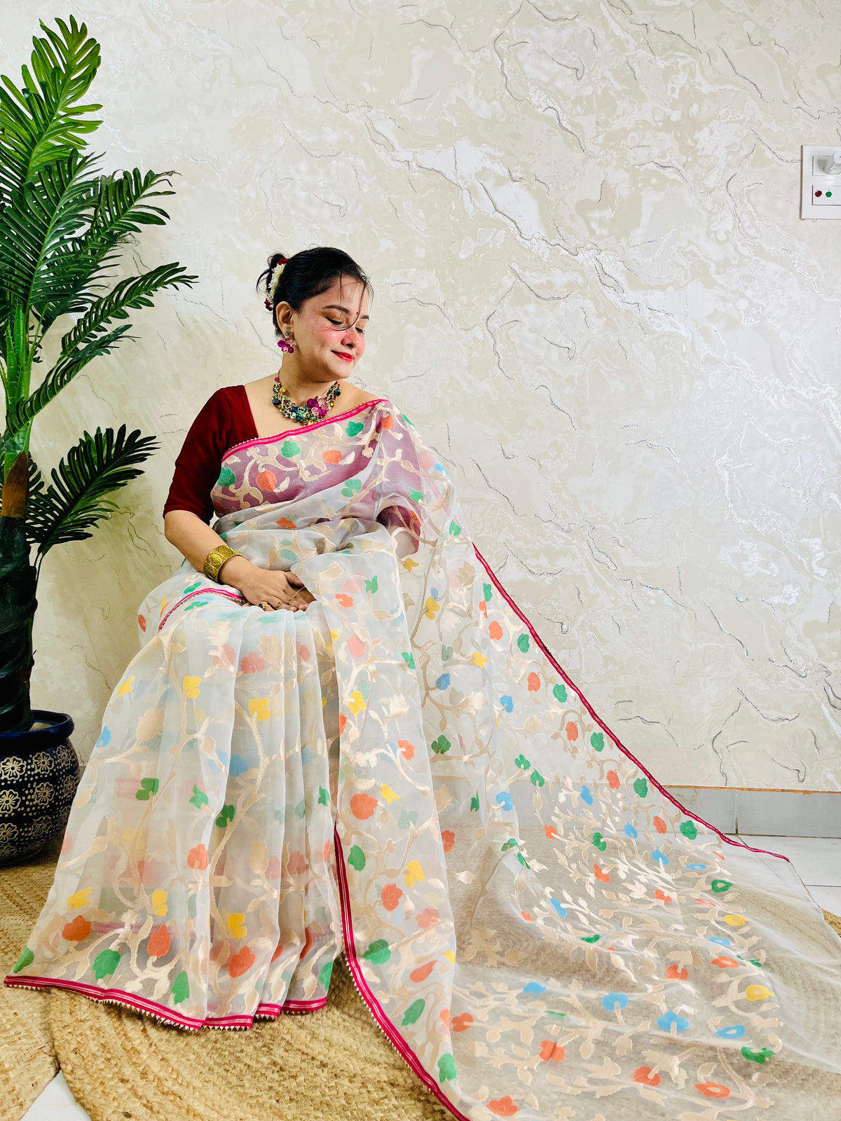 A woman wearing a white saree with colorful floral prints and a deep red blouse, seated indoors against a textured cream wall with a potted plant on the left.