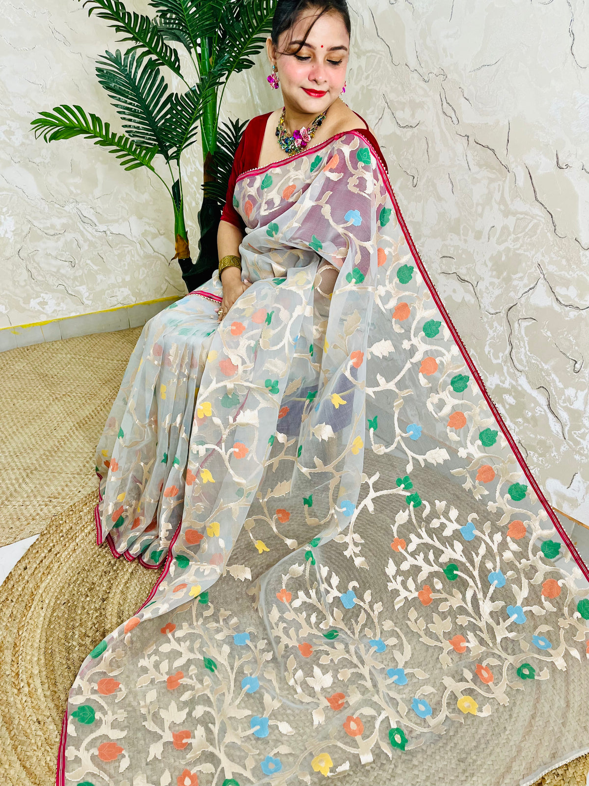 A woman wearing a sheer beige saree with colorful floral embroidery and a red border, seated on a woven mat against a textured wall with a potted plant.