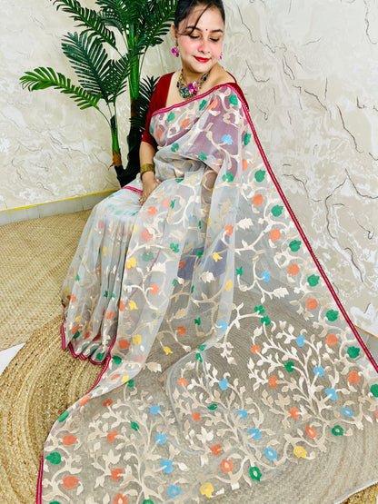 A woman wearing a sheer beige saree with colorful floral embroidery and a red border, seated on a woven mat against a textured wall with a potted plant.