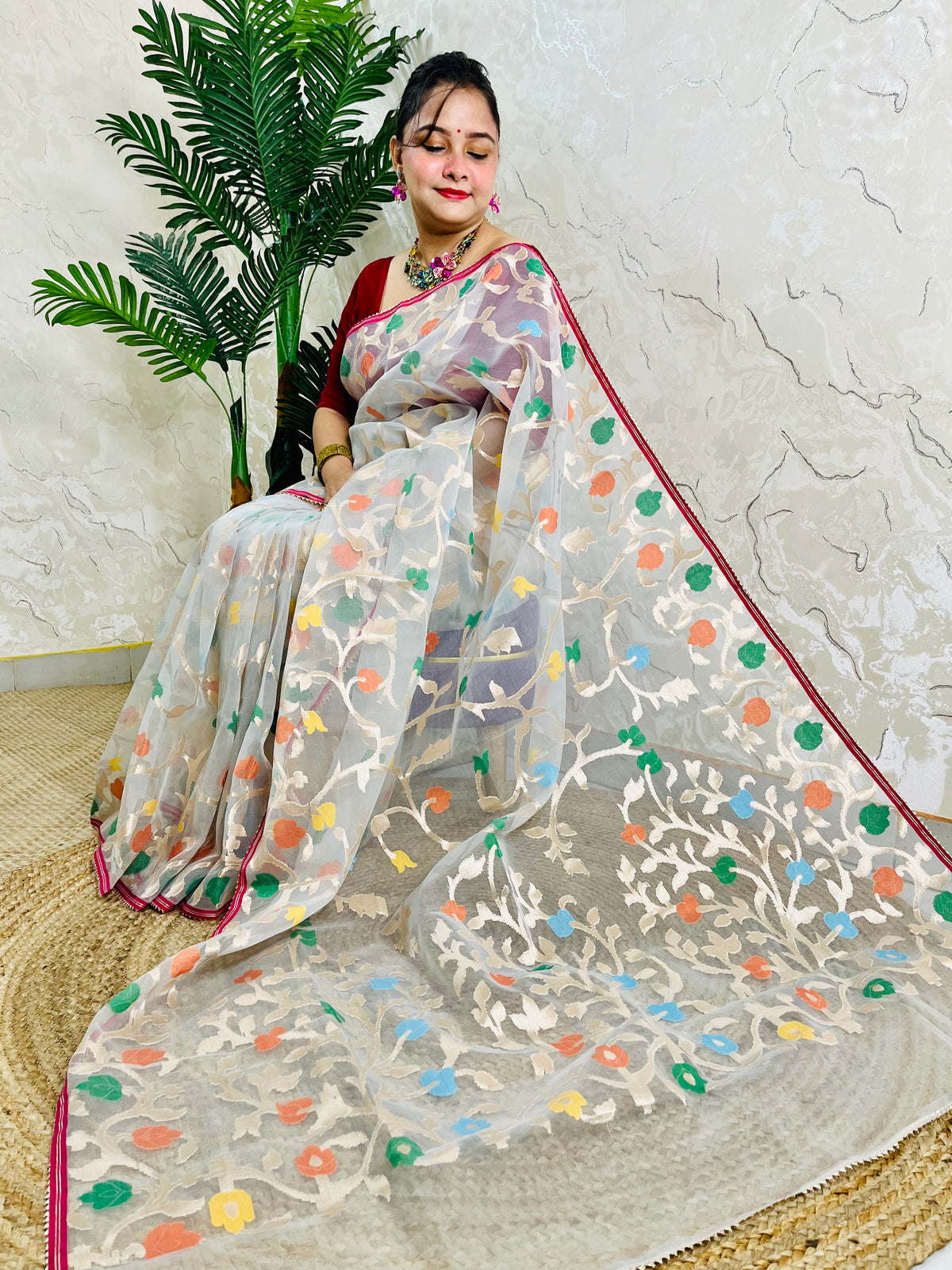 A woman seated indoors wearing a sheer off-white saree with multicolored floral motifs and a red border, paired with a red blouse. A potted palm and textured wall are visible in the background.
