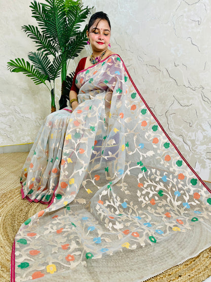 A woman seated indoors wearing a sheer off-white saree with multicolored floral motifs and a red border, paired with a red blouse. A potted palm and textured wall are visible in the background.