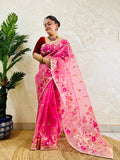 A woman wearing a pink floral saree with a red blouse, standing indoors next to a potted plant against a textured beige wall.