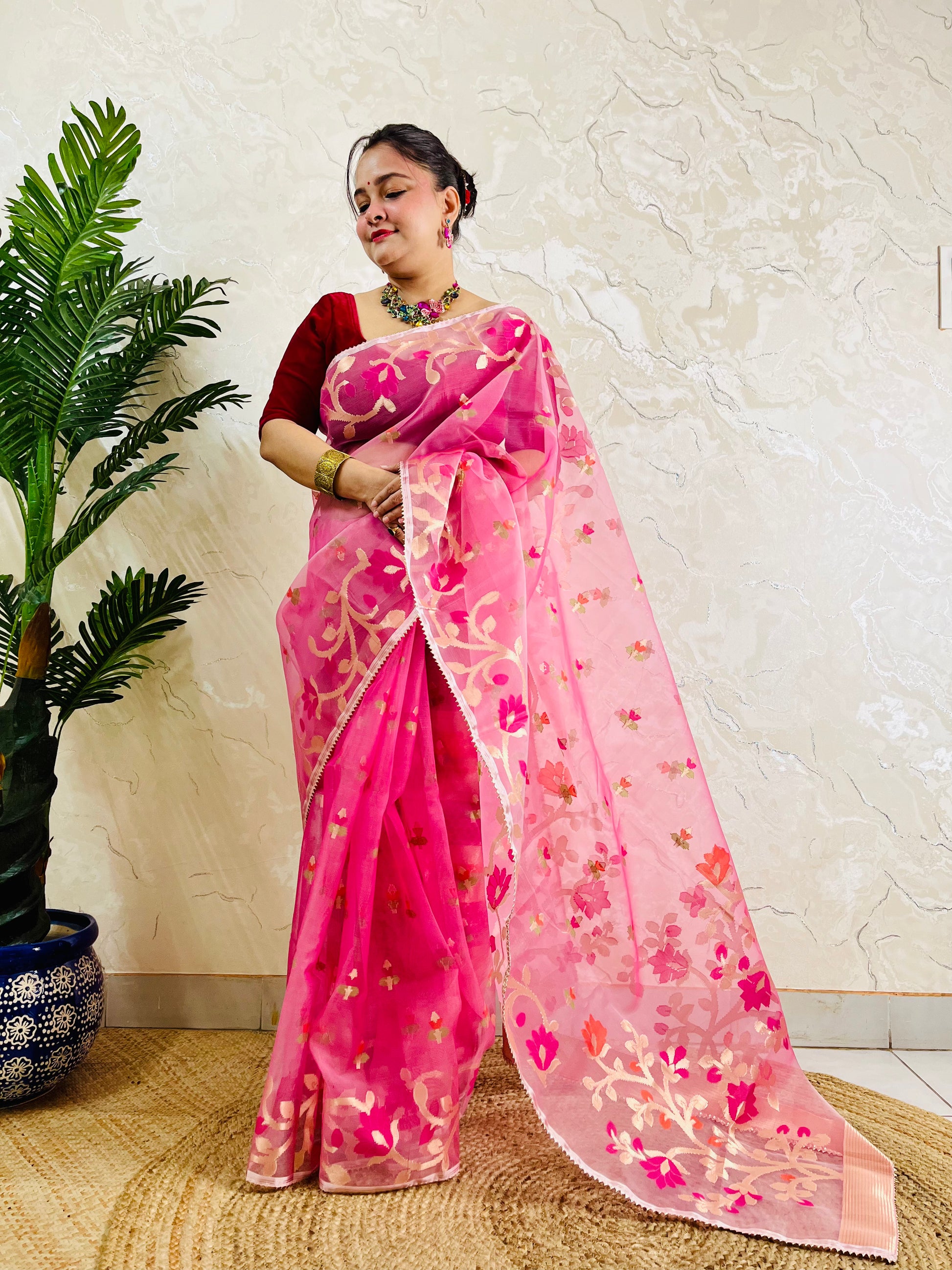 A woman wearing a pink floral saree with a red blouse, standing indoors next to a potted plant against a textured beige wall.