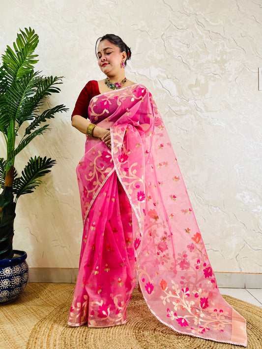 A woman wearing a pink floral saree with a red blouse, standing indoors next to a potted plant against a textured beige wall.
