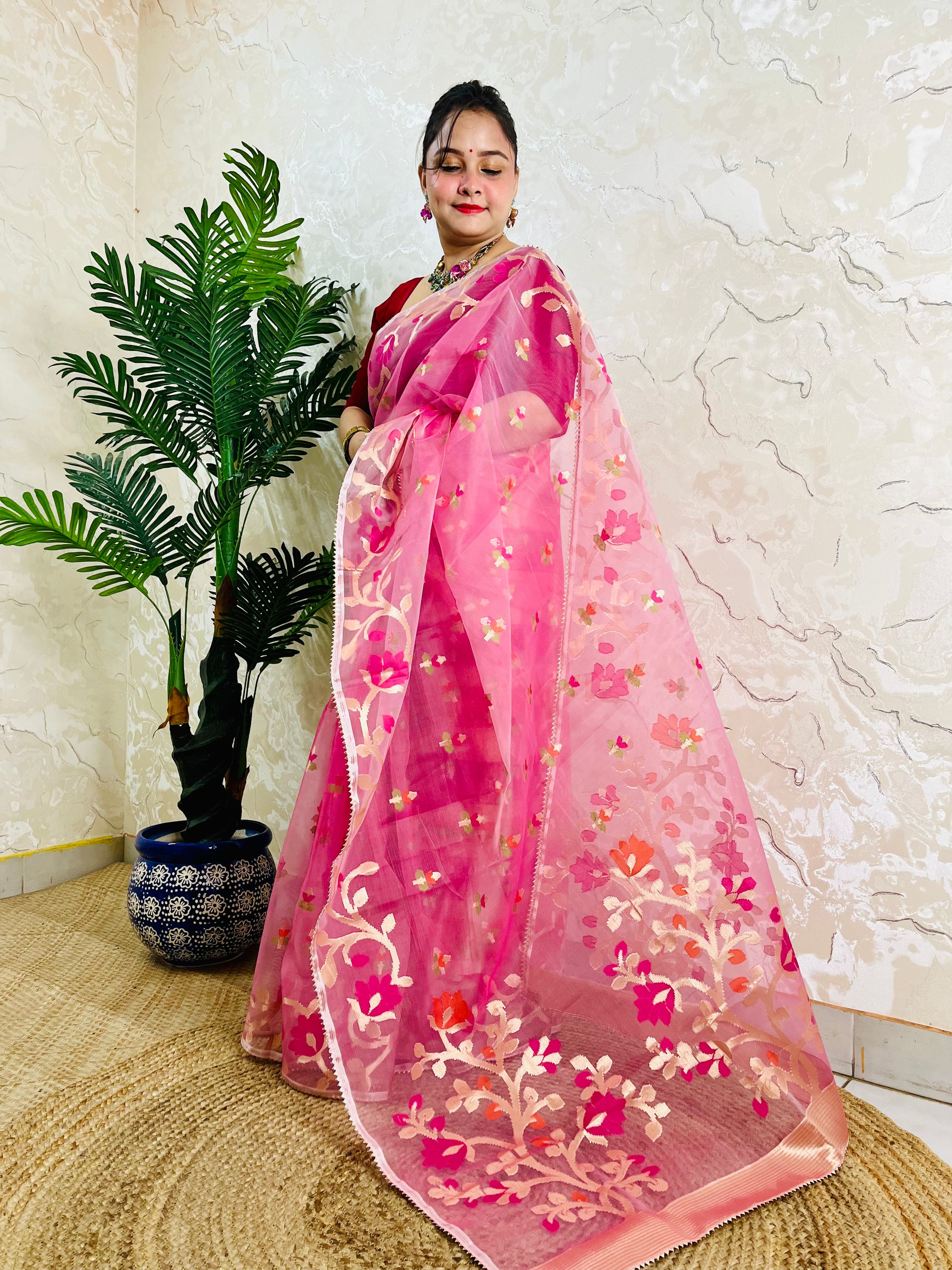 A woman wearing a pink embroidered saree with floral patterns, standing indoors next to a potted palm tree against a textured cream wall.