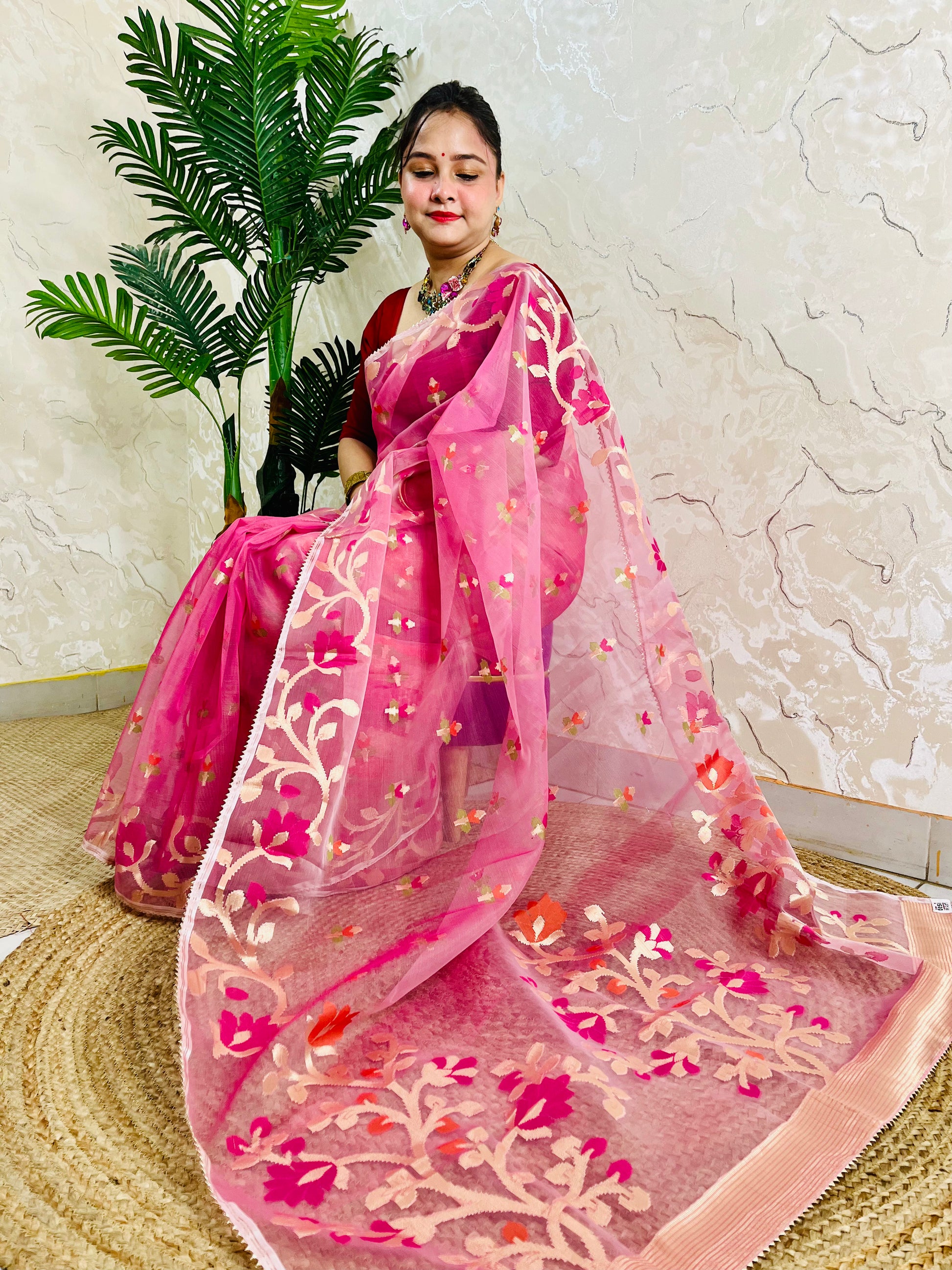 A woman models a vibrant pink saree with a matching sheer dupatta. The saree features intricate floral embroidery in pink, gold, and orange, and is styled with a red blouse.