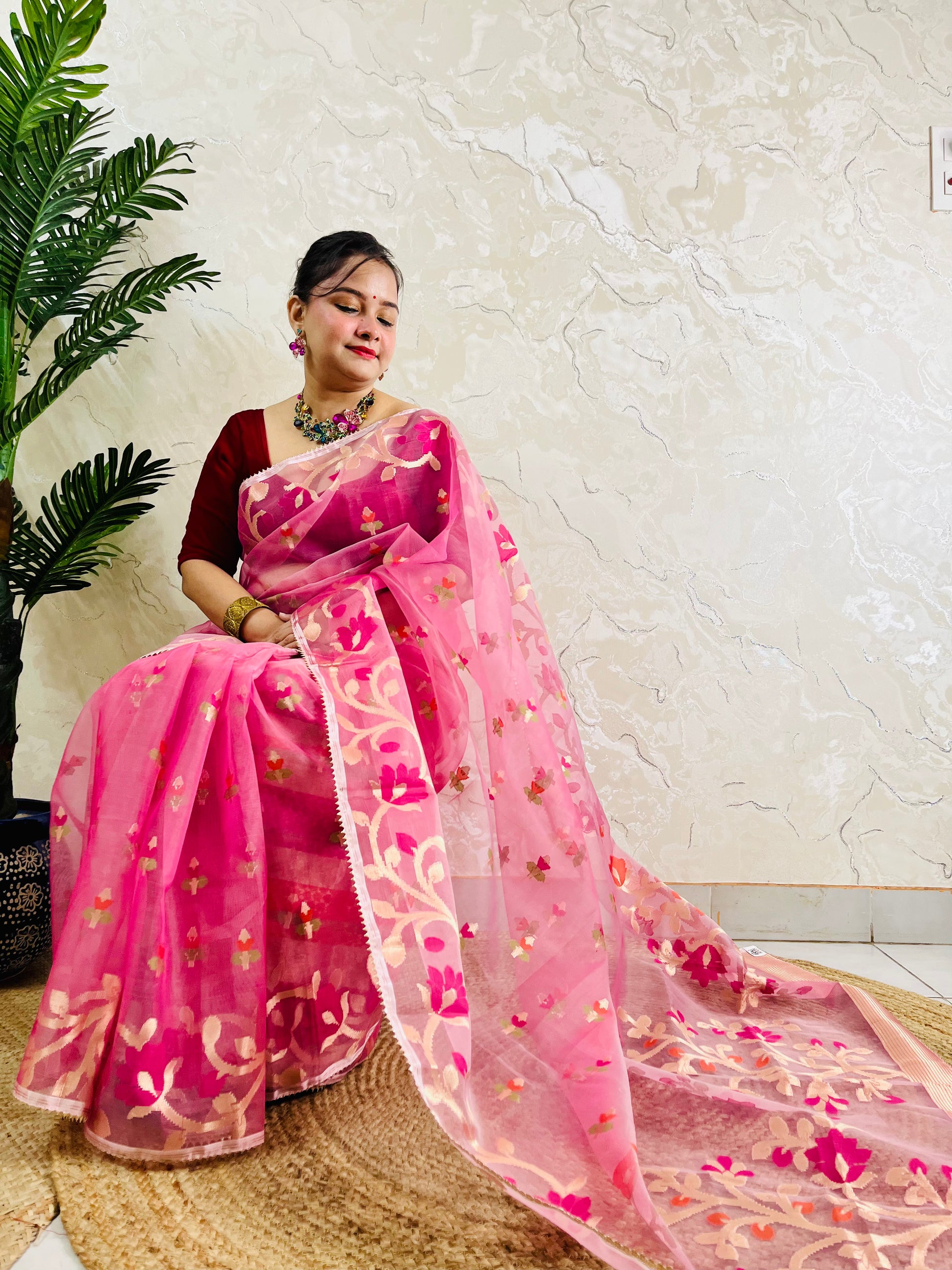 A woman wearing a pink floral embroidered saree with a deep red blouse, seated on a woven mat against a textured beige wall with a green plant on the left.