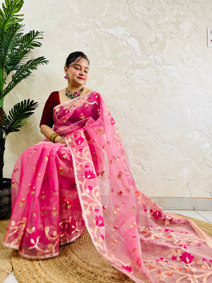 A woman wearing a pink floral embroidered saree with a deep red blouse, seated on a woven mat against a textured beige wall with a green plant on the left.