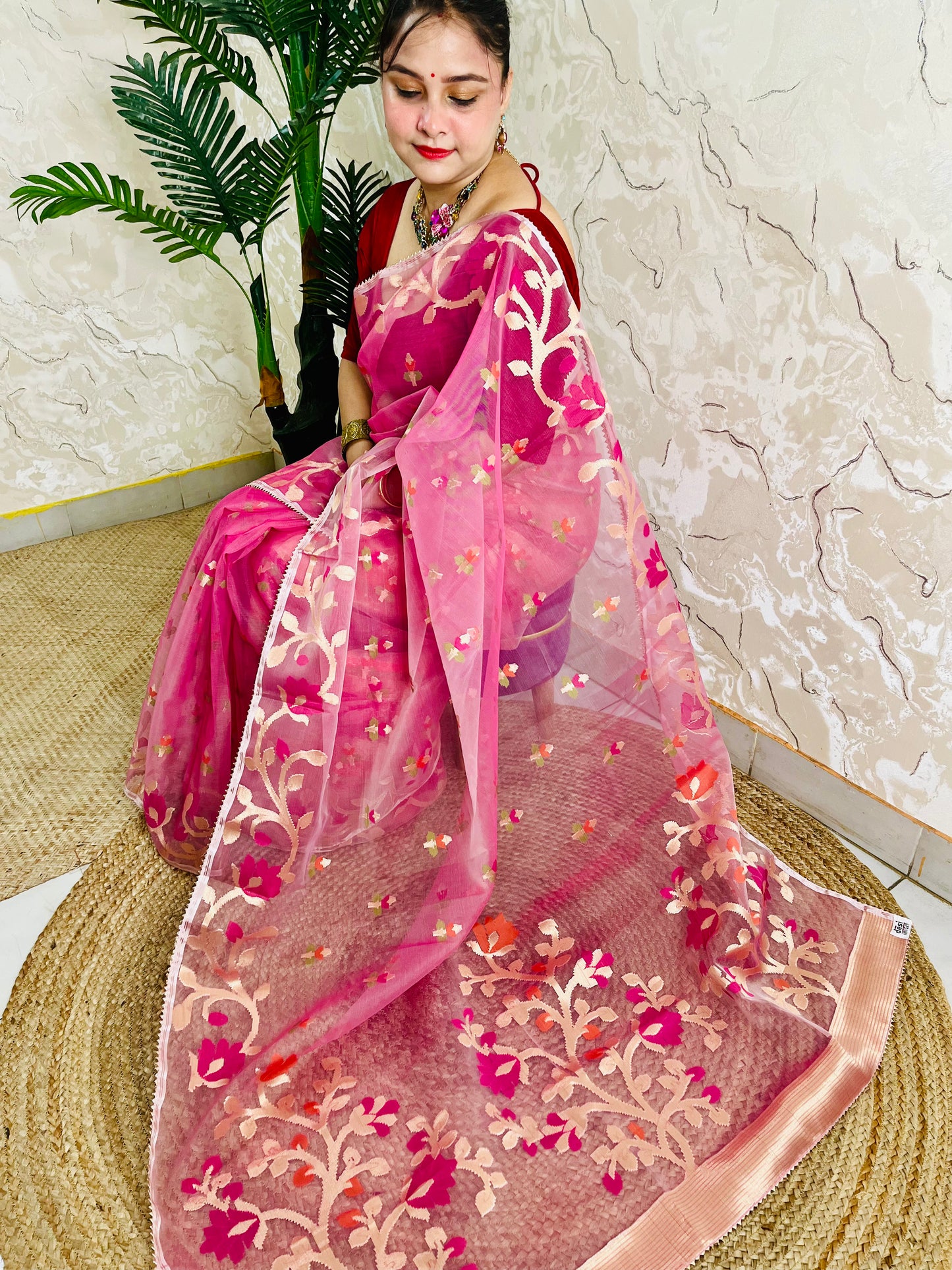 A woman wearing a bright pink embroidered saree with a matching sheer dupatta, seated on a woven mat against a textured beige wall and potted plant.