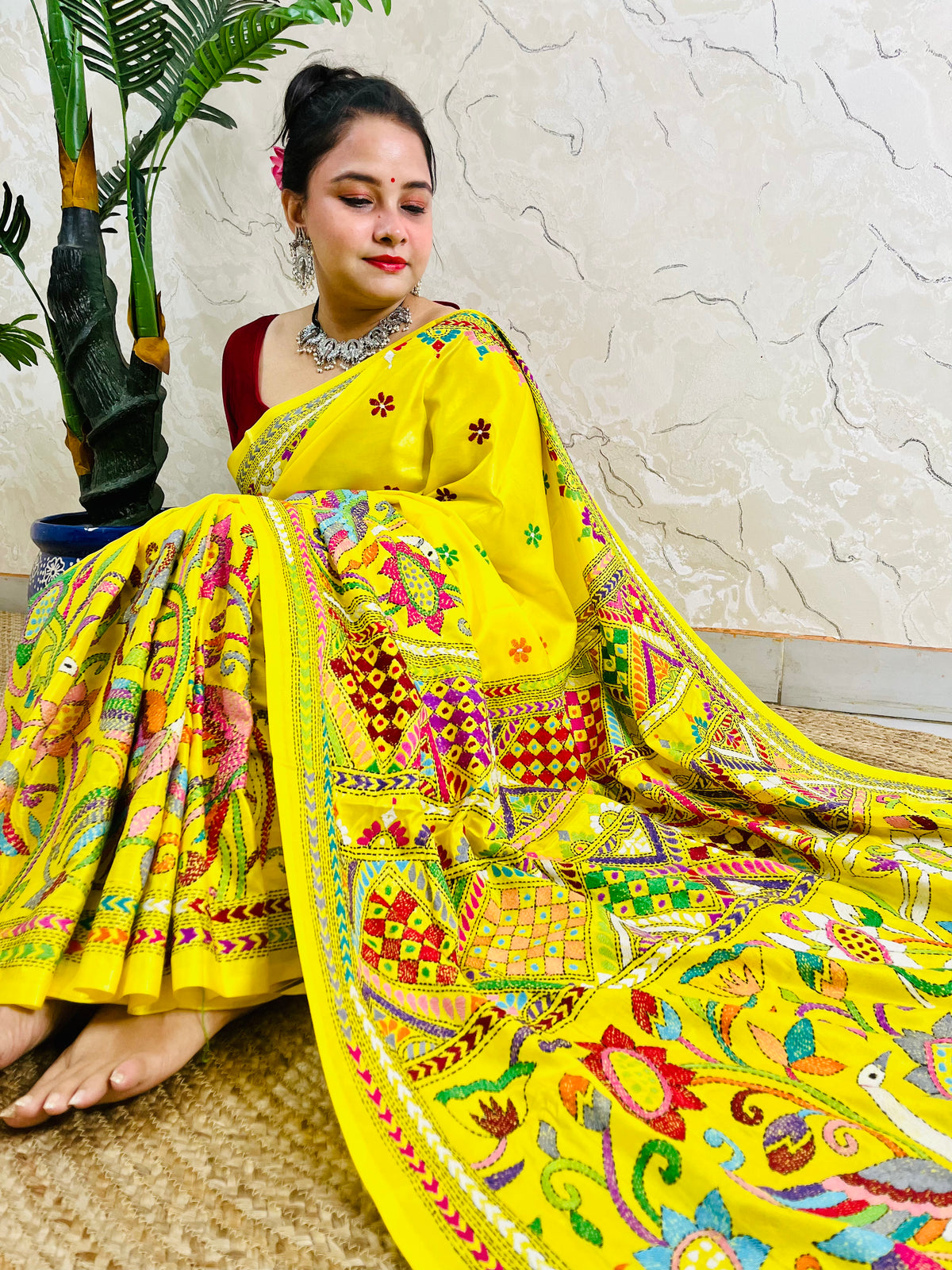 Woman sitting gracefully while draped in the vibrant sunny yellow Nakshi Katha saree, showcasing the densely embroidered, multi-colored border and pallu