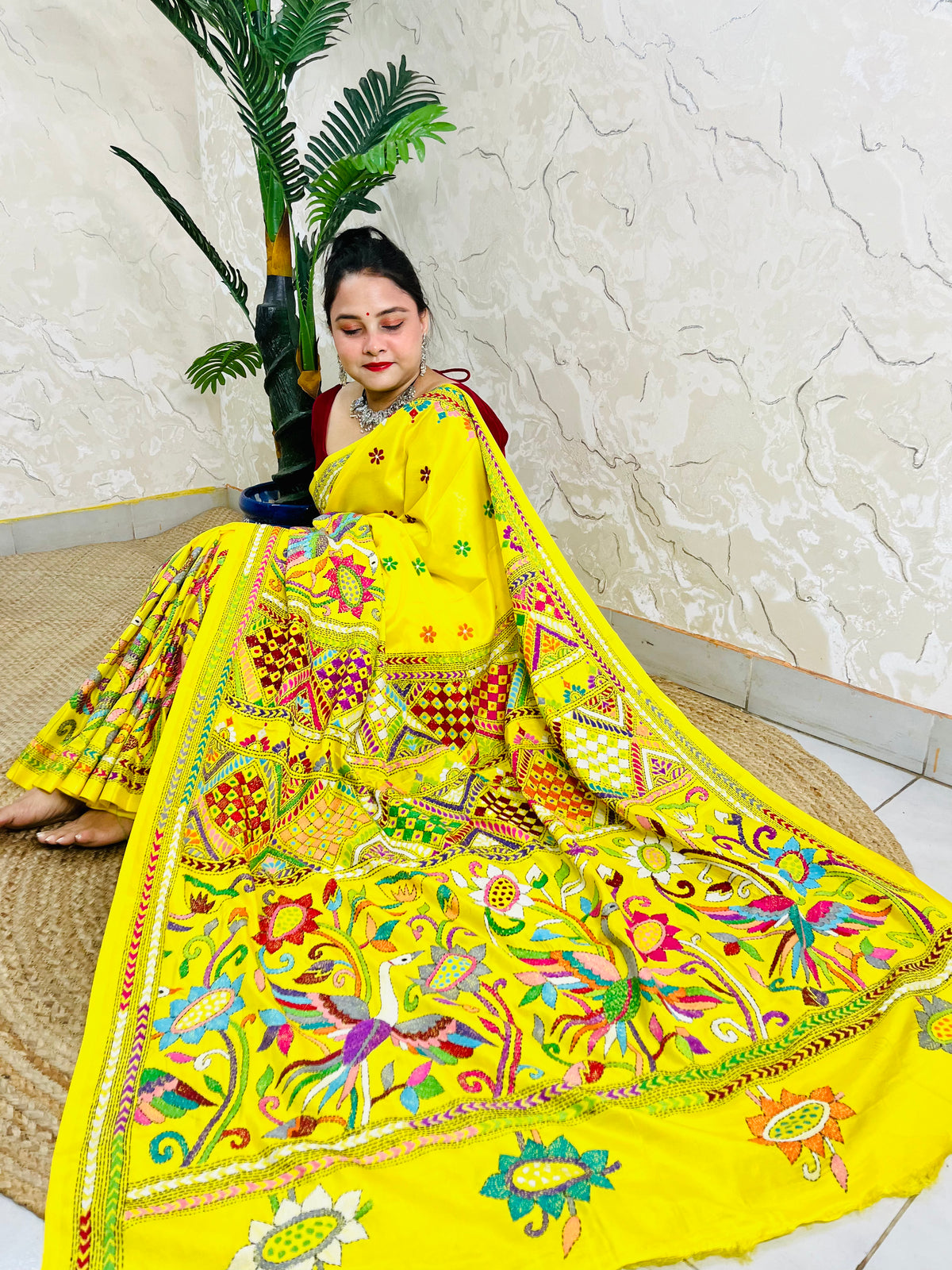 Woman sitting gracefully while draped in the vibrant sunny yellow Nakshi Katha saree, showcasing the densely embroidered, multi-colored border and pallu