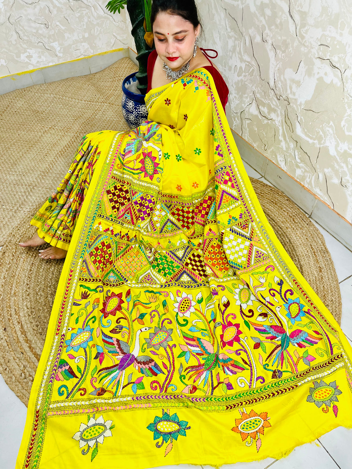 Woman sitting gracefully while draped in the vibrant sunny yellow Nakshi Katha saree, showcasing the densely embroidered, multi-colored border and pallu