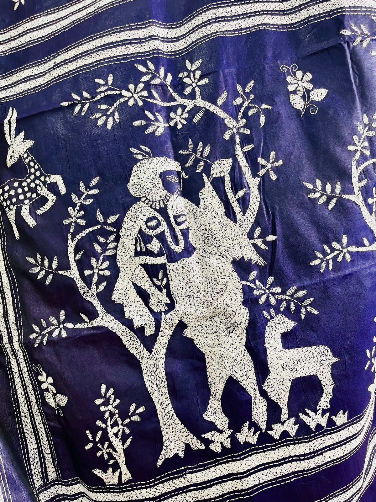 Close-up shot of white running thread Katha stitch embroidery depicting a Santhal flute player, trees, and deer on navy silk.