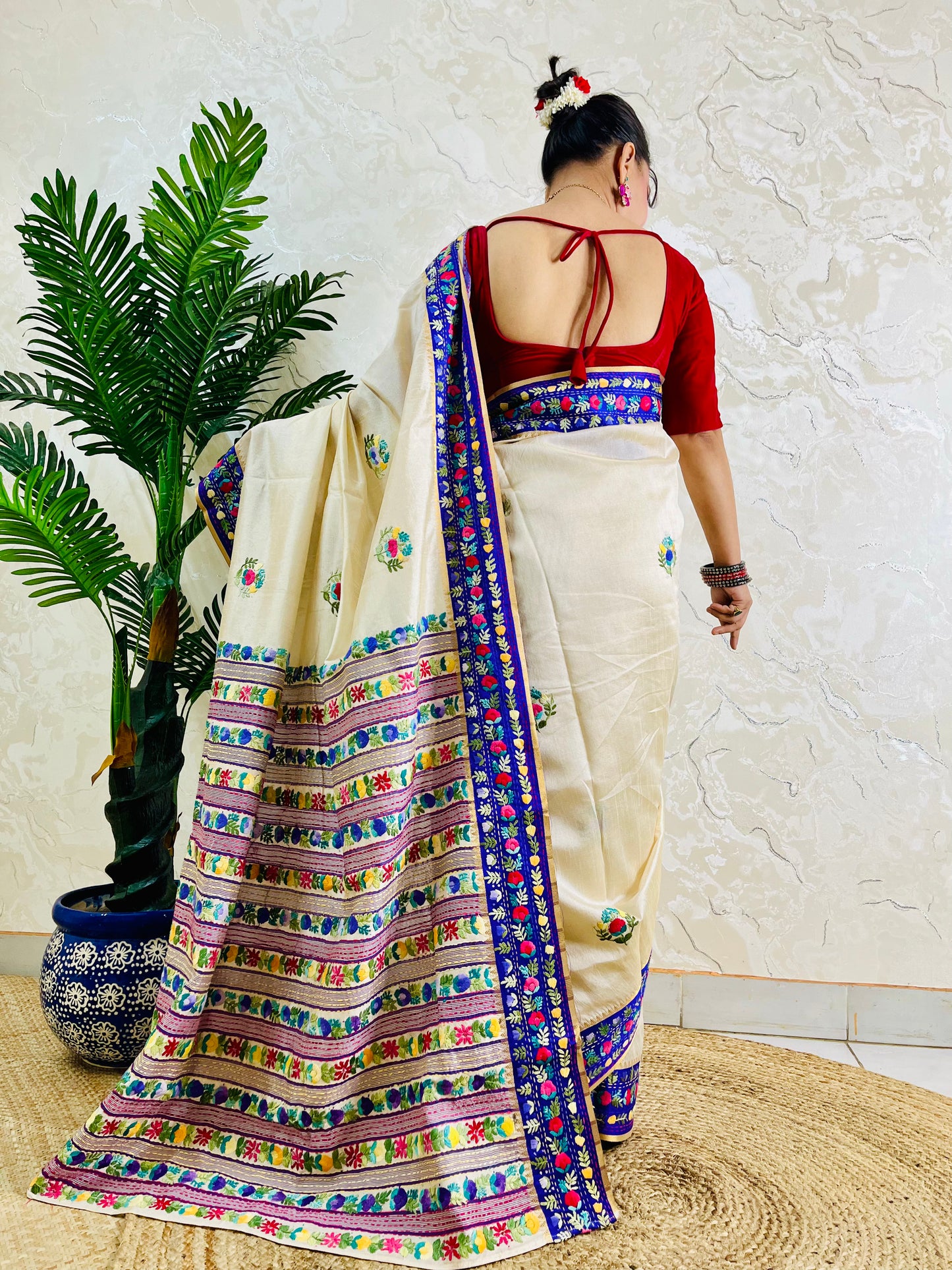 A woman shown from behind wearing a cream-colored saree with a red backless blouse, featuring a highly detailed multicolor border and floral motifs, standing in front of a textured wall next to a potted plant.