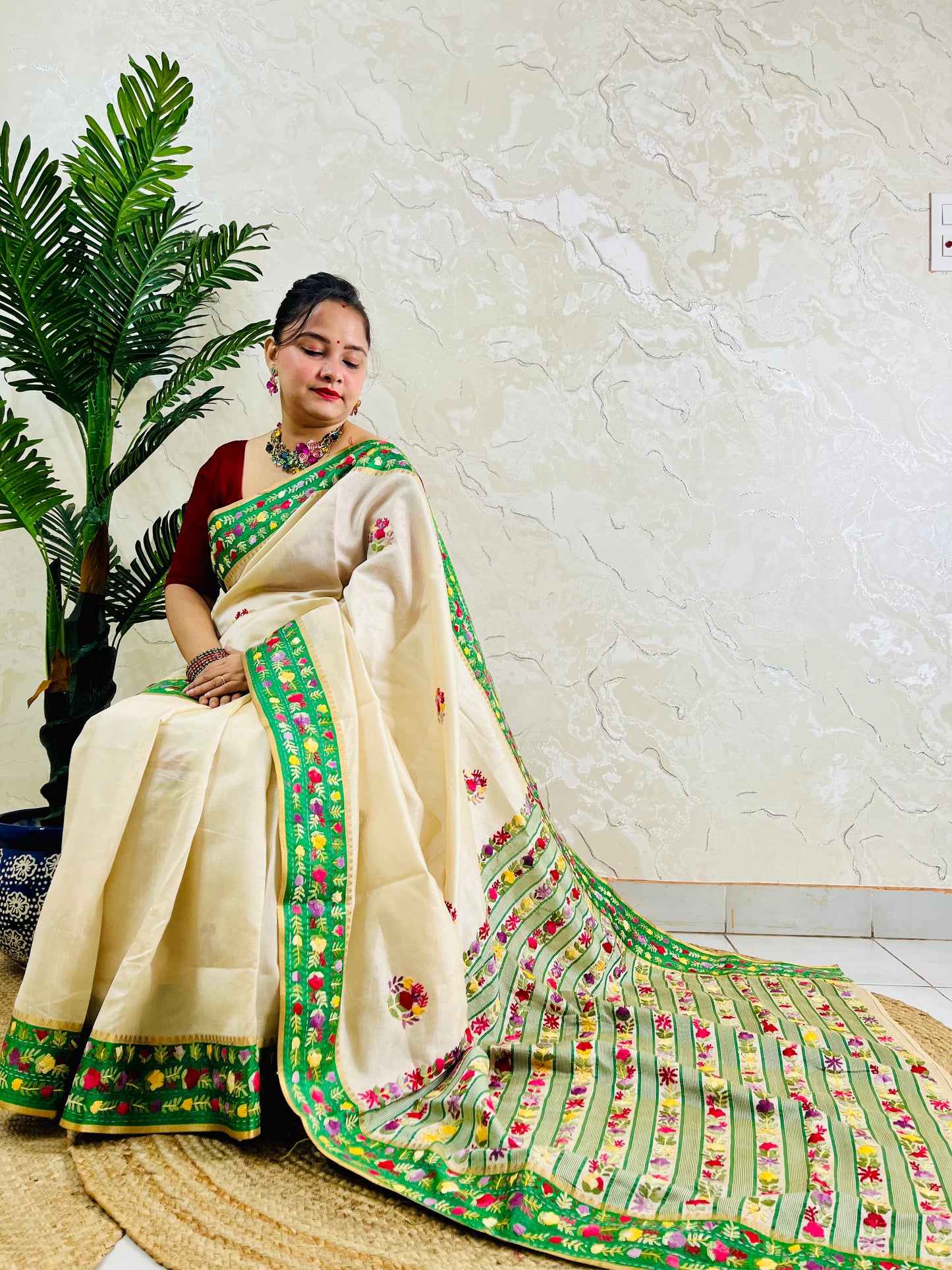 Side sitting view of Parsi Gara handwoven ivory Semi Muga Silk saree with emerald green velvet border and full multicolour floral garden pallu with woven stripe body — Desi Aadat