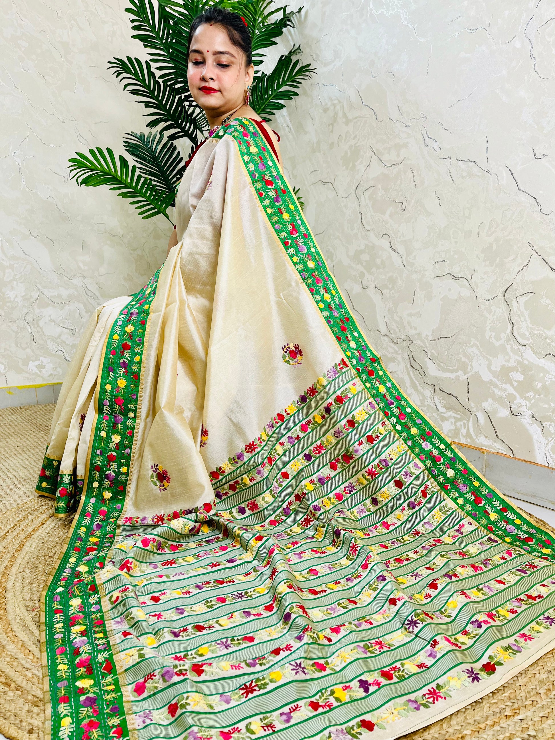 Side sitting view of Parsi Gara handwoven ivory Semi Muga Silk saree with emerald green velvet border and full multicolour floral garden pallu with woven stripe body — Desi Aadat