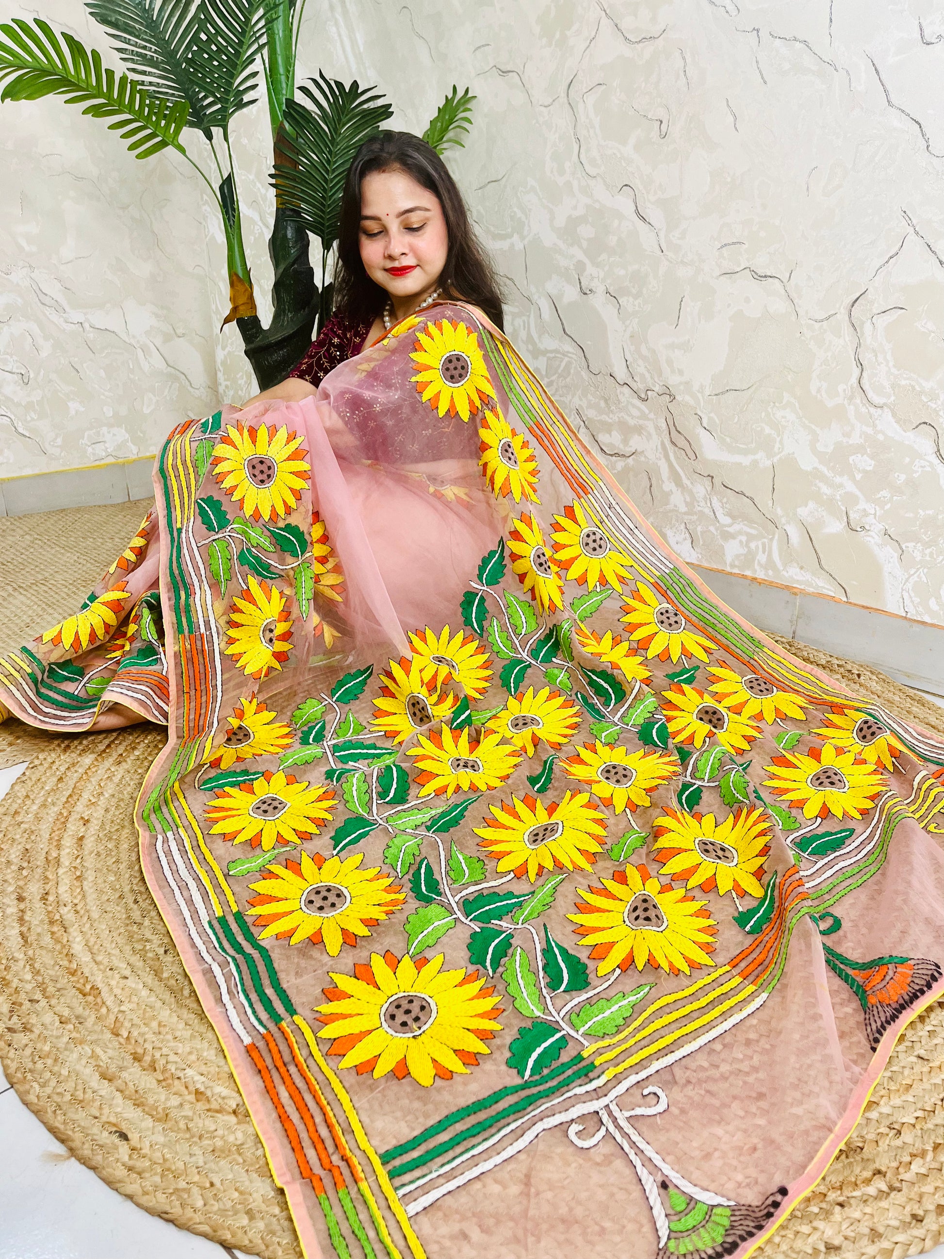 Handmade sunflower Kantha stitch blush pink organza saree with large golden yellow sunflower blooms and multicolour stripe border — angled sitting drape view, Desi Aadat