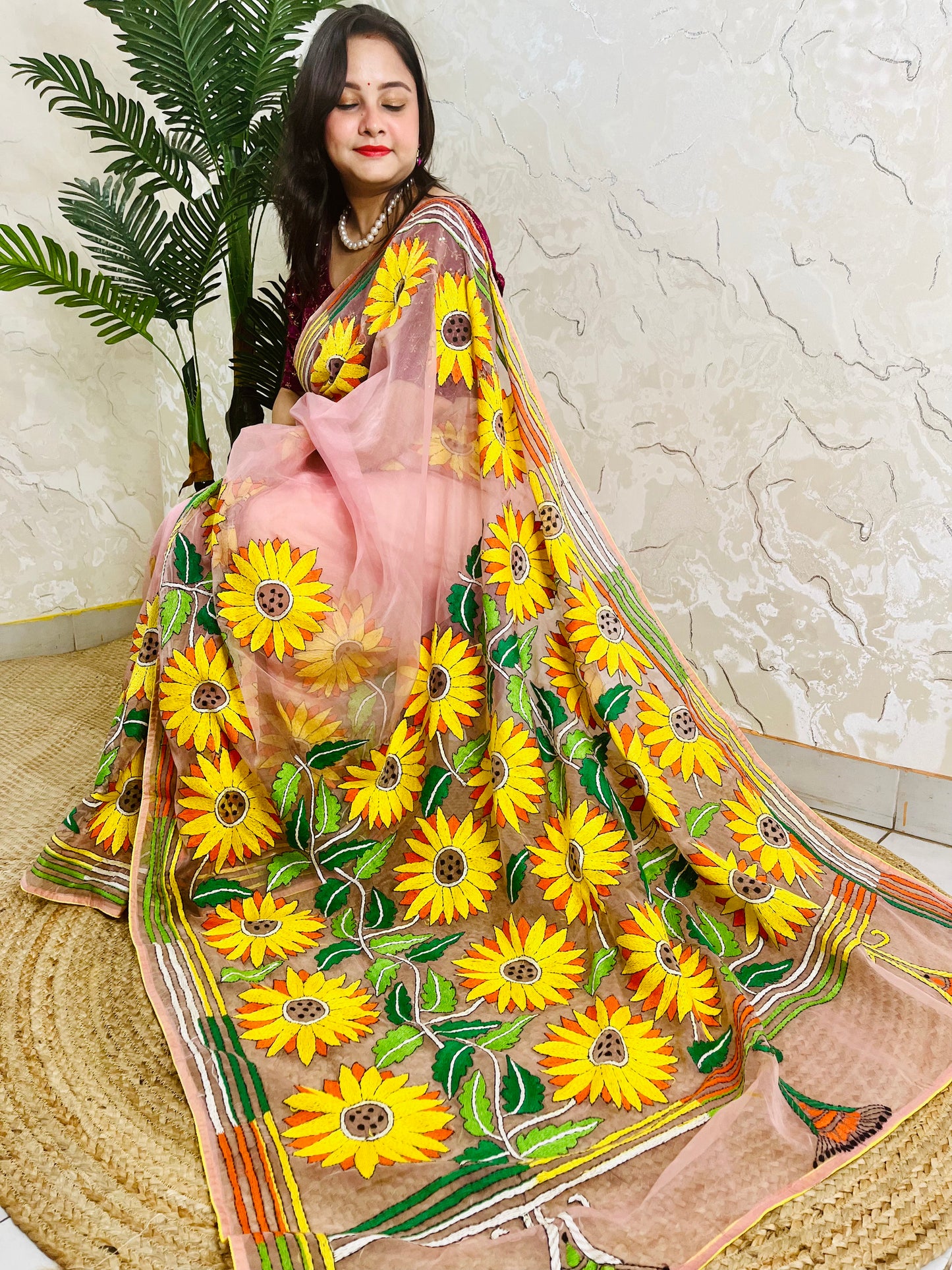 Handmade sunflower Kantha stitch blush pink organza saree with large golden yellow sunflower blooms and multicolour stripe border — angled sitting drape view, Desi Aadat