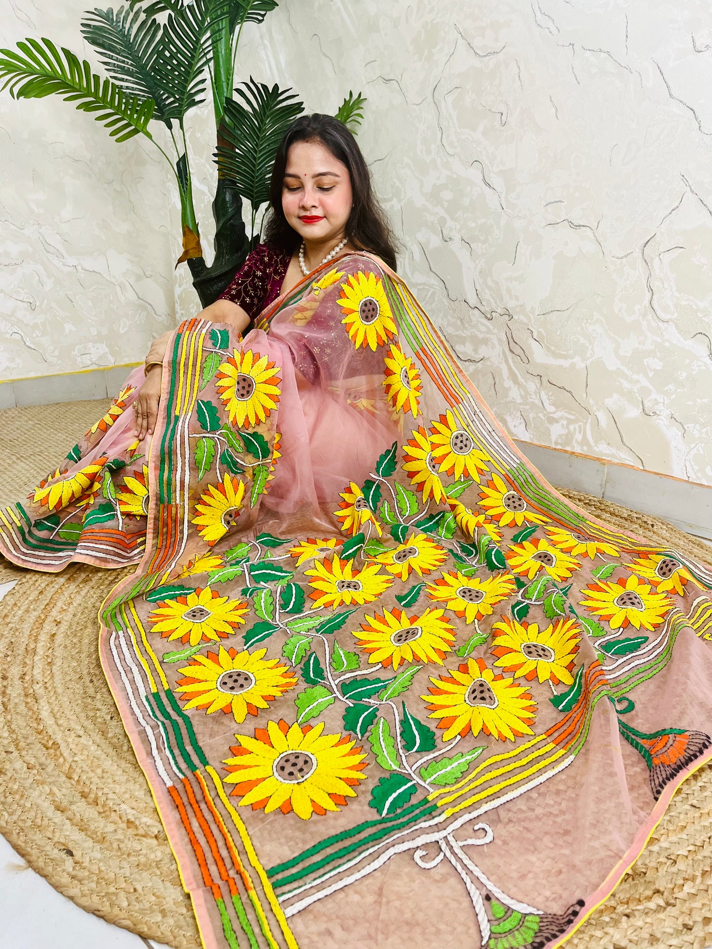 Handmade sunflower Kantha stitch blush pink organza saree with large golden yellow sunflower blooms and multicolour stripe border — angled sitting drape view, Desi Aadat