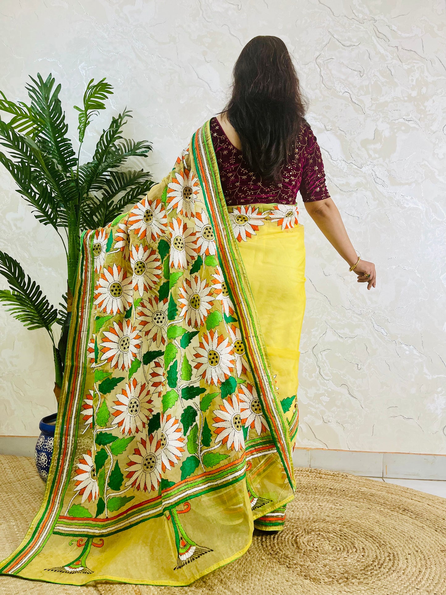 Handmade sunflower Kantha stitch sunny yellow organza saree with white daisy blooms, orange petals, green leaves, and multicolour stripe border — standing back view, Desi Aadat