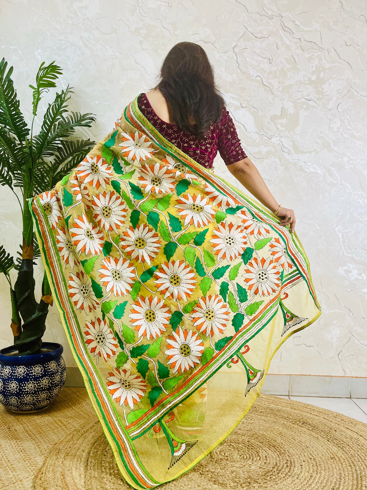 Handmade sunflower Kantha stitch sunny yellow organza saree with white daisy blooms, orange petals, green leaves, and multicolour stripe border — standing back view, Desi Aadat