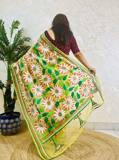 Handmade sunflower Kantha stitch sunny yellow organza saree with white daisy blooms, orange petals, green leaves, and multicolour stripe border — standing back view, Desi Aadat