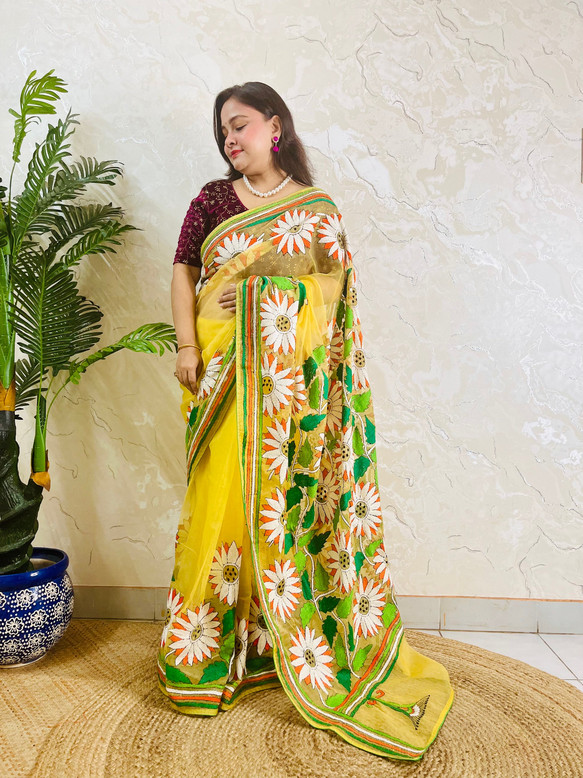 Handmade sunflower Kantha stitch sunny yellow organza saree with white daisy blooms, orange petals, green leaves, and multicolour stripe border — standing view, Desi Aadat