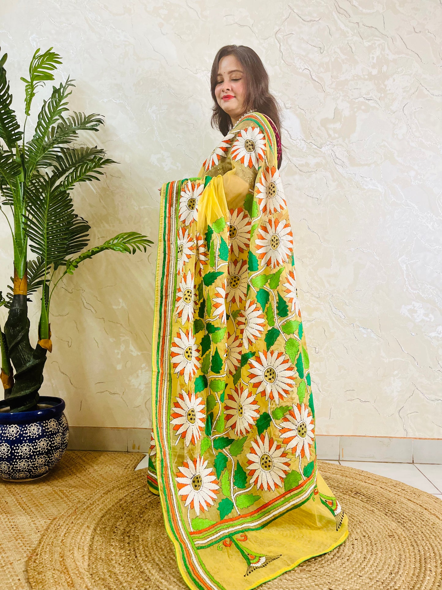 Handmade sunflower Kantha stitch sunny yellow organza saree with white daisy blooms, orange petals, green leaves, and multicolour stripe border — standing view, Desi Aadat