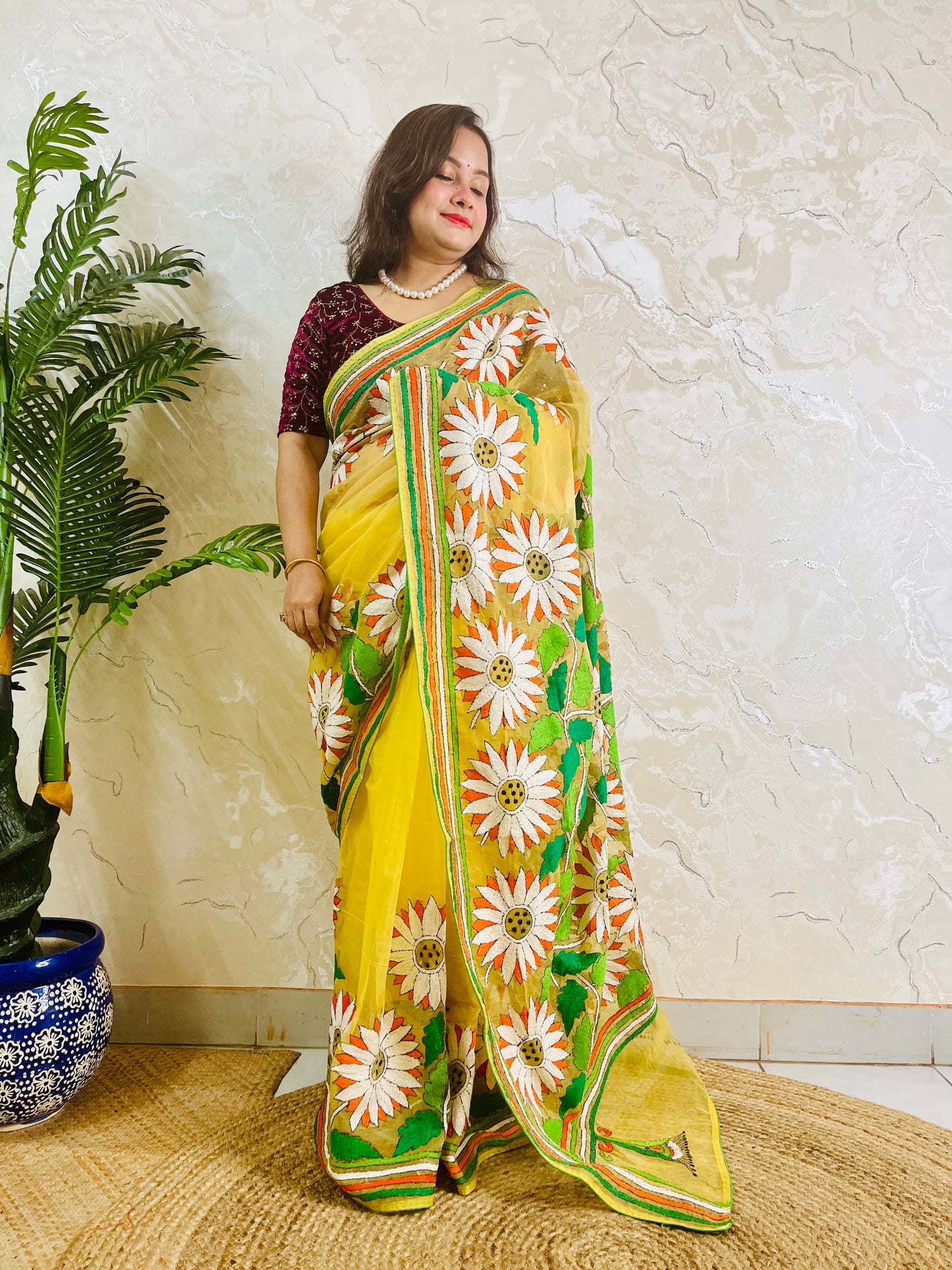 Handmade sunflower Kantha stitch sunny yellow organza saree with white daisy blooms, orange petals, green leaves, and multicolour stripe border — standing view, Desi Aadat