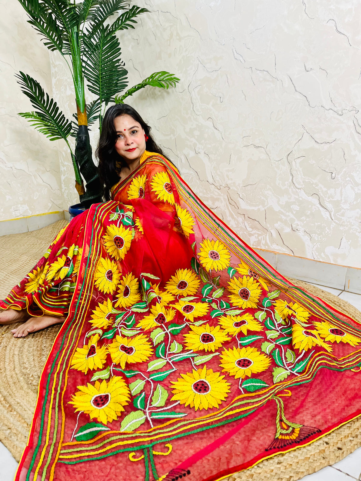 Woman seated in crimson red sunflower Kantha stitch saree, angled view