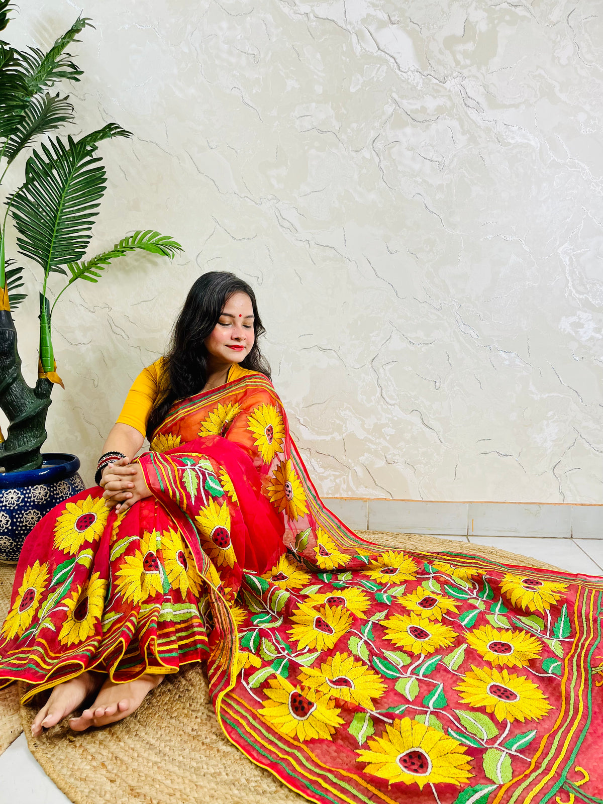 Woman seated in crimson red sunflower Kantha stitch saree, angled view