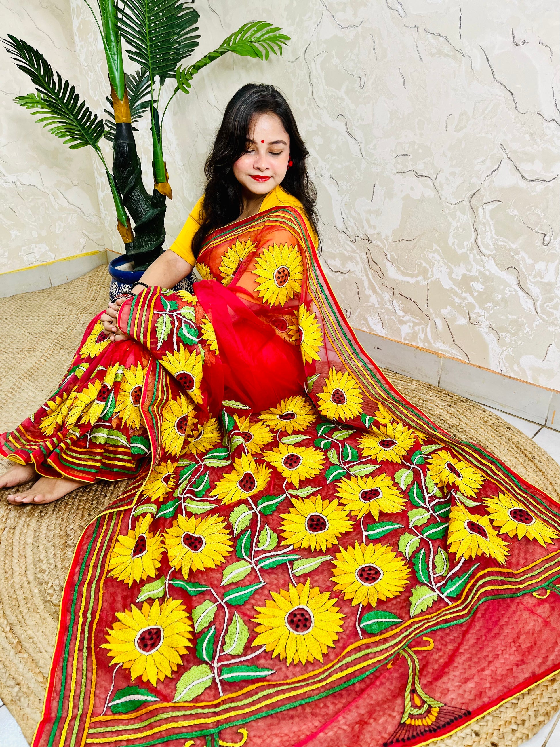 Woman seated in crimson red sunflower Kantha stitch saree, angled view